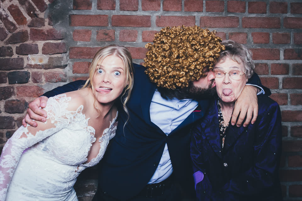 Fotobox-Bild: Braut, ein Mann mit großer Lockenperücke vor dem Gesicht und eine ältere Dame, die die Zunge herausstreckt.