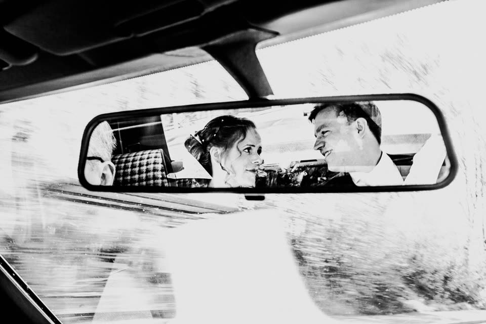 Schwarzweißfoto durch den Rückspiegel: Braut und Bräutigam lächeln sich im Auto an.