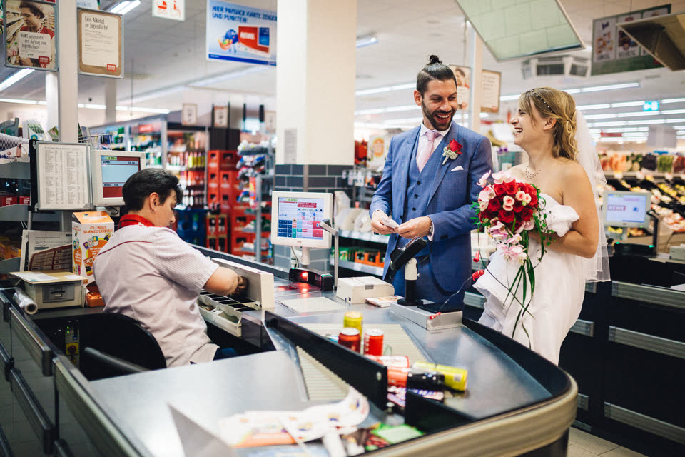 Brautpaar mit Brautstrauß an der Supermarktkasse, die Kassiererin kassiert.